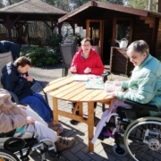 Vier Frauen, zwei davon im Rollstuhl, sitzen vor einem Gartenhäuschen an einem runden Holztisch in der Sonne. Sie basteln. Im Hintergrund limks von Ihnen bauen die beiden Männer den Jenga-Turm auf.