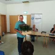 Manfred Groth steht mit einer Stiege mit Blumen in beiden Händen. Er wendet sich den Mitgliedern zu.