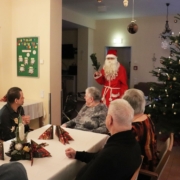Der Weihnachtsmann mit rotem Kapuzenmantel, weißem Bart und Rute in der rechten Hand betritt den Raum. Rechts von ihm steht ein großer Weihnachtsbaum. Im Vordergrund sind einige Bewohner zu sehen.