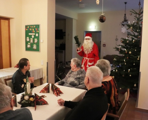 Der Weihnachtsmann mit rotem Kapuzenmantel, weißem Bart und Rute in der rechten Hand betritt den Raum. Rechts von ihm steht ein großer Weihnachtsbaum. Im Vordergrund sind einige Bewohner zu sehen.