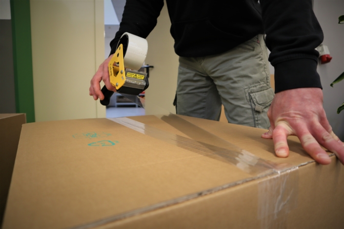 Ein Beschäftigter sichert einen großen braunen Karton mit breitem, durchsichtigem Klebeband.