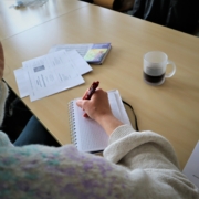 Eine Teilnehmerin macht sich in einem kleinen Ringbuch Notizen. Auf dem Tisch liegen verschiedene Unterlagen. Auch ein Tasse Kaffee steht dabei.