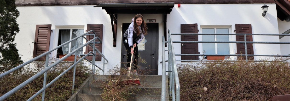 Eine junge Frau fegt vor dem Giebel eines 'Fachwerkhauses Gestrüpp eine Betontreppe herunter. Diese hat links und rechts ein graues Geländer.