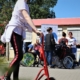 Eine Besucherin ist mit einem roten Roller auf dem Schulhof unterwegs. Dahinter stehen mehrere Kinder und Jugendliche im Rollstuhl am Stand der Schulbegleitung.